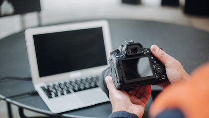 Professional photographer checking digital photos on camera screen, laptop nearby with blank display, focused on post production workflow