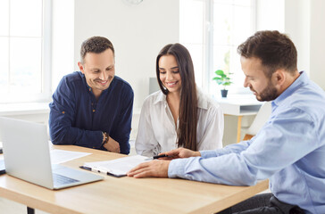 Professional real estate agent sitting at desk with happy clients, finalizing details of their new home purchase. Young cheerful family couple preparing to sign contract agreement at realtor office.