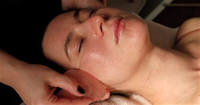 Young woman receiving facial massage with gua sha tool in beauty salon