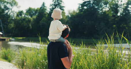 Caring father carries his son on his shoulder, wearing a hat, by a pond watching surfing competitions in the midst of nature.