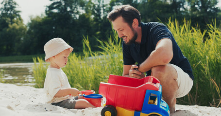 Boy with a hat on plays in the sandbox on the beach, pouring sand into a bucket, with his dad...