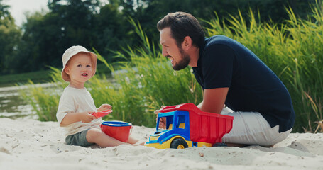 Dad and son enjoy their time outdoors on the beach, as the boy with a hat plays in the sandbox, pouring sand into a bucket and receiving a high-five.