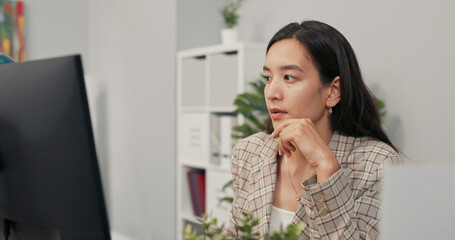 A focused pretty woman with long brown hair wearing a checkered jacket sits at desk in office in...