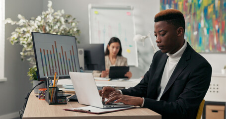 A smiling dark-skinned boy spends morning at work in front of computer screen, tapping fingers on laptop keyboard, filling out official and company applications, comparing correctness on two monitors