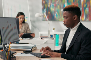A focused dark-skinned man in suit spends time at work in front of office computer, completing charts on monitor, transcribing data onto pieces of paper, signing documents, completing applications