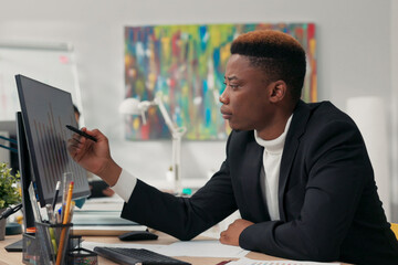 Handsome dark-skinned man, corporate office worker spends morning in front of computer, points at screen, counts, ponders, checks correctness of graphs, completes data