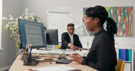 The manager, boss, office director with dark skin dressed in black suit sits behind desk prepares documents for mailing asks assistant to pass envelopes girl fulfills request and returns to her duties