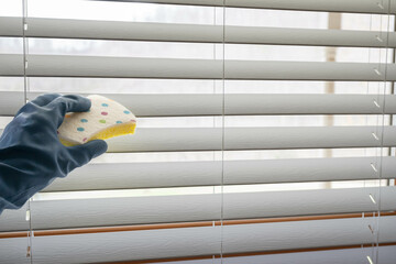 Hand with dark blue glove cleaning window coverings with a polka-dot sponge.