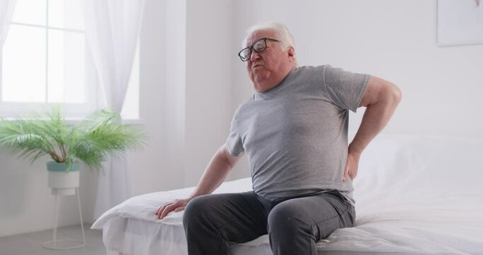Upset senior man suffering from lower back pain, trying to get up from bed. Gray haired old pensioner experiencing intense discomfort from backaches and lumbar pain, feeling strong spasm in his spine.
