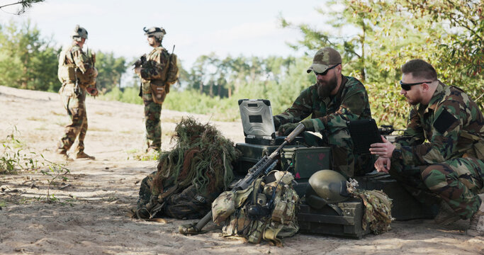 Soldiers in full combat gear, including helmets and backpacks, prepare for battle at the training ground, reviewing strategy, entering data, and holding a tablet while discussing their approach.