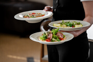 A waiter elegantly serves three plates of gourmet food, showcasing fresh salads and ingredients. The setting is upscale, creating a warm dining atmosphere.