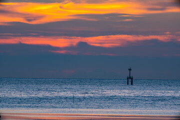 A striking seascape capturing the vivid hues of a sunset sky blending orange, red, and purple over the calm ocean. A navigation tower stands against the horizon, adding depth and dramatic scene.  .