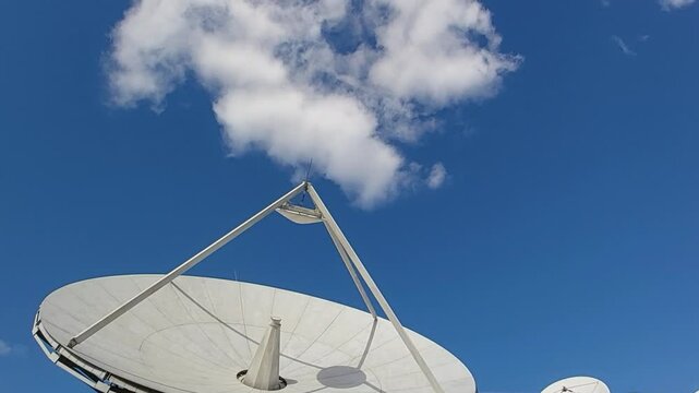White parabolic antenna pointing to the blue sky with some white clouds passing by