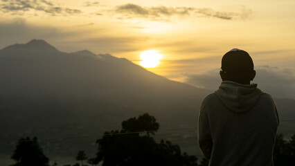the silhouette of a man wearing a hoodie and a cap, standing while facing a breathtaking sunset behind the mountains. The sky is filled with golden hues blended with gradients of orange and soft gray