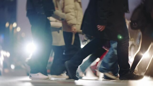 Low angle close-up view people crowd feet various shoes steps busy street. Many pedestrians walking crowded crosswalk backlit with bright car headlights. Evening rush hour legs asphalt zebra crossing