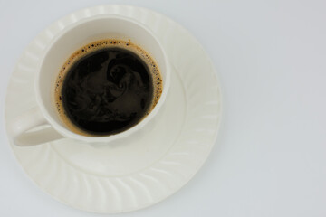 A white mug of coffee stands on a white saucer, black aromatic coffee with foam around the edges on a white background with space for text is poured into the mug. 