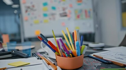 Colorful pens and markers sit in a container on a designer's desk, surrounded by sketches and notes, in a modern office environment with a whiteboard filled with ideas in the background