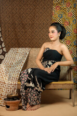 Fototapeta premium Indonesian woman in a black batik dress, sitting elegantly on a wooden bench. She exudes grace with a traditional hairstyle, vintage makeup, and cultural charm in a classic setting.