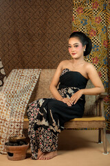 Fototapeta premium Indonesian woman in a black batik dress, sitting elegantly on a wooden bench. She exudes grace with a traditional hairstyle, vintage makeup, and cultural charm in a classic setting.