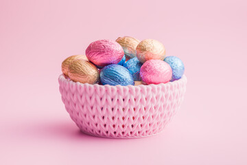 Chocolate easter eggs wrapped in multi colored foil in bowl on pink background.