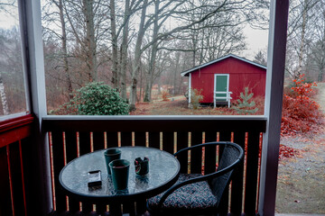 Skåne, Sweden-January 12, 2025: Swedish countryside with traditional red wooden house and cozy porch. Forest cabin. A cozy porch with a round glass table, two green coffee mugs, a pack of cigarettes. 