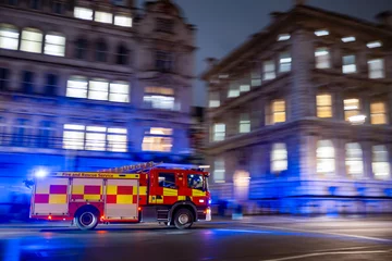 Fototapeten Fast red firetruck moving on emergency call in London city center with tall bouldings around. lights on and firefighters inside. motion effect. panning effect. fire brigade UK  © JairoMZ
