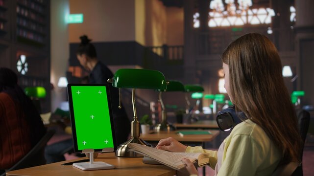 Schoolgirl reading a book next to copy space on tablet at the library desk, using academic database for research. Completing school assignments and developing her educational skills. Camera A.
