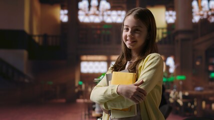 Portrait of sweet preadolescent carrying her books and class notes in the library, preparing for an...