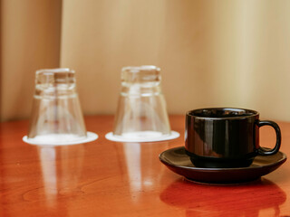 A black coffee or tea cup sits on a red table with two glasses. Hotel room for two persons.