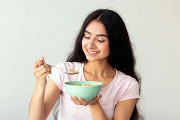 A young Indian woman smiles while holding a bowl of cereal, enjoying a nutritious breakfast at...