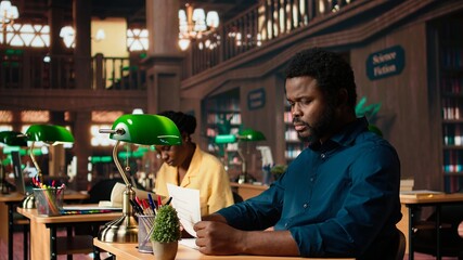 Black university student feeling anxious and struggling with learning, trying to memorize relevant information from course notes at the library. Male pupil studying to pass exams. Camera A.