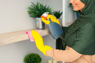 A young Muslim woman in a headscarf applies cleaner to a shelf while using a cloth. She smiles as...