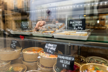 Close-up of a bakery display case filled with take-away meals and desserts. A hand arranges a tray of pastries inside. Glass reflection, price tags, shop counter facing the street, blurred background