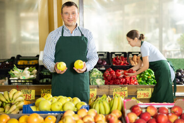 Man seller of garden stuff department store is replenishing showcase with fruits, puts apples on...
