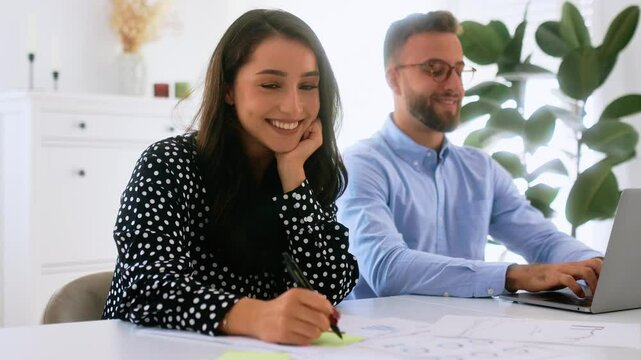 Happy charming young arabian or caucasian brunette woman writing on a sticky note at a desk and smiles at the camera while a man types on a laptop in the background, showcasing teamwork and creativity