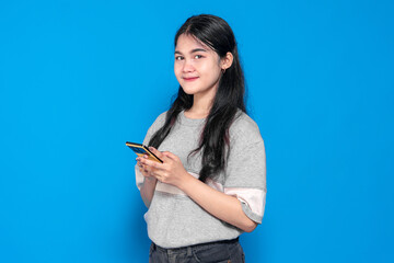 Naklejka premium A young Indonesian woman with long black hair, wearing a casual gray t-shirt, smiling while holding a smartphone against a bright blue background. Confident and relaxed pose.