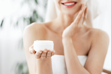 Unrecognizable woman in towels applying cream on face after shower, holding jar with cream, copy...