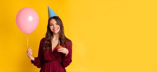 A young woman in a burgundy dress smiles while holding a pink balloon and a piece of cake. She wears a festive party hat, embodying the cheerful spirit of a birthday celebration, copy space