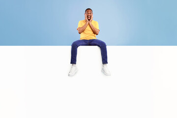 A joyful African American man sits against a vibrant backdrop, his hands on his cheeks in excitement. This lifestyle shot captures a moment of pure happiness in an isolated setting.