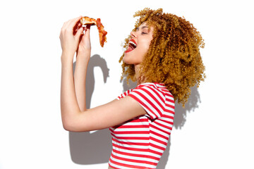 Young woman with curly hair enjoying a slice of pizza, wearing a striped shirt, expressing...