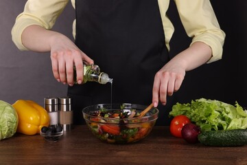 Woman pouring oil onto tasty salad at wooden table against dark background, closeup