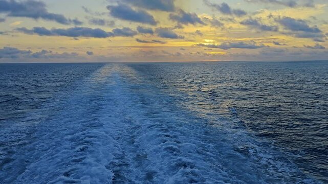 Golden sunrise peeks over the horizon setting clouds aglow aft of ship on calm ocean with ship's wake trailing off to the distant horizon 