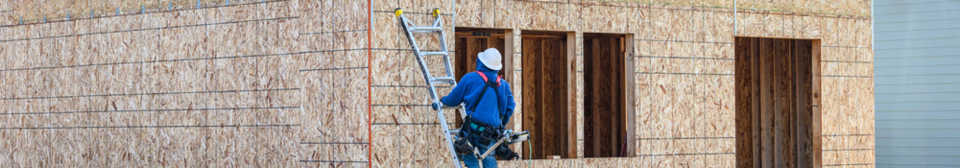 Fototapeta premium Carpenter in hard hat and toolbelt on ladder against exterior OSB (oriented strand board) wall, using hammer to attach metal tie plate, new home construction site, housing development 