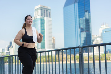 Portrait of young woman jogs along a railing by a waterfront, surrounded by modern skyscrapers on a clear day. Asian plus size woman