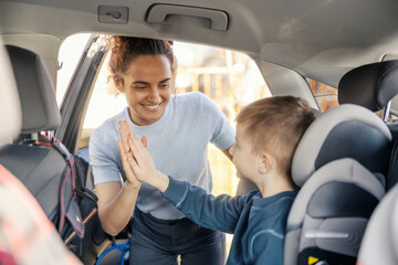 Portrait of a young mom giving high five to her son while he is sitting in child seat in a car.