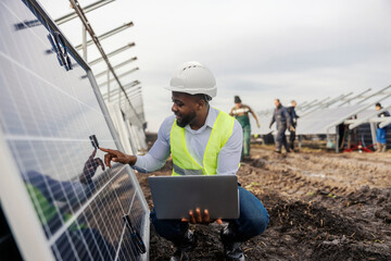 Smiling black engineer with laptop at solar farm inspecting quality of solar panels.