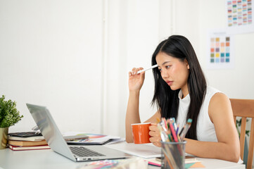 An asian woman is thinking thoughtfully while looking at laptop with a mug and a stylus in each hand