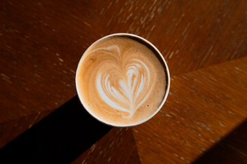 A cup of latte with heart-shaped latte art placed on a wooden table in warm lighting.