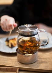 A glass teapot with loose leaf tea sits on a warmer at a cozy cafe setting.