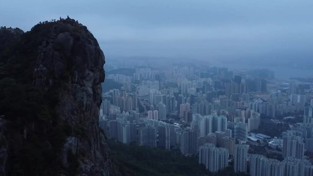 Gorgeous aerial drone footage of the city Hong Kong in China during a misty foggy or smog covered dusk or dawn day with a large mountain ricky cliff in the foreground. Tall building visible in city.
